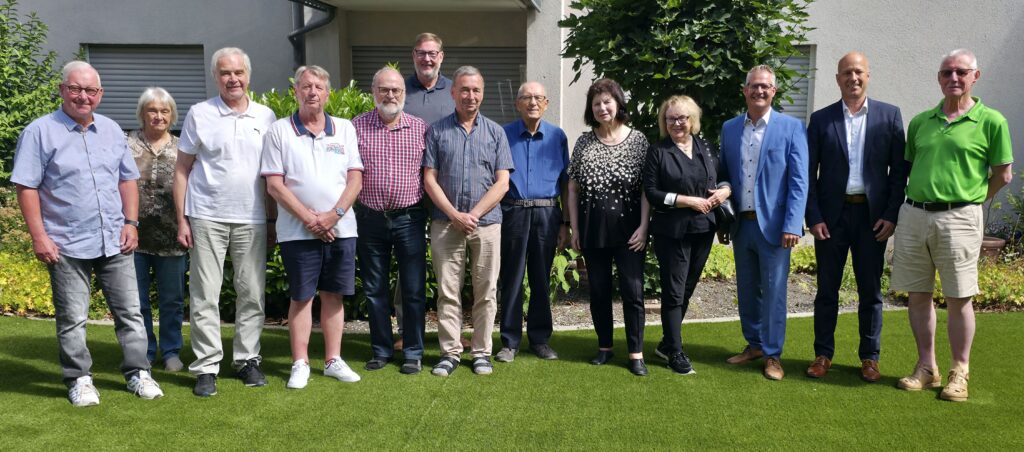 Ein Dutzend Seniorinnen und Senioren in Freizeitkleidung stehen bei schönem Wetter im Halbkreis auf einem Rasen, blicken in die Kamera  und lächeln.